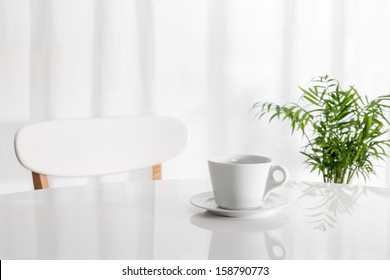 White Cup On The Kitchen Table, With Green Plant In The Background.