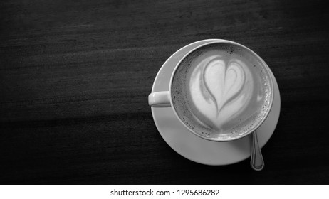 White cup and saucer with a heart shape foam latte art on a wooden table in black and white, monochrome.