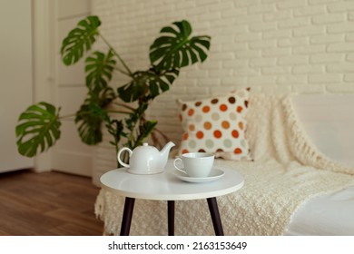 A White Cup With A Saucer And A Teapot On A Small Table Near The Sofa