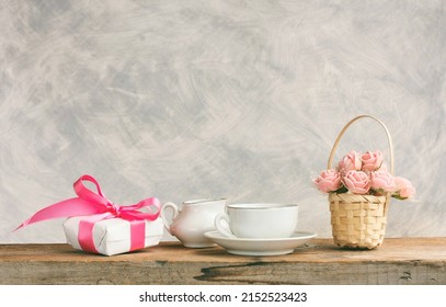 white cup with tea, gift box, basket with roses on wooden table, congratulation concept