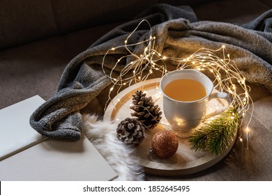 A White Cup Of Tea On A Wooden Serving Tray With Pine Cones And In The Background A Fairy Lights On A Sofa In The Living Room. Wool Blanket, Christmas, Advent.