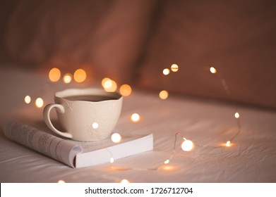 White cup of tea staying on open book with glowing Christmas lights at background. Good morning. Breakfast. Stay at home.  - Powered by Shutterstock