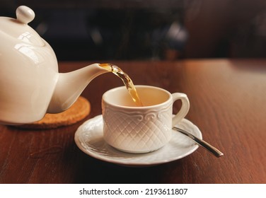 White cup with teapot. The hand holding the teapot pours tea into a pot standing on saucer in soft focus on naturally blurred background. Coffee, tea house, bokeh lights. The concept of a cozy - Powered by Shutterstock