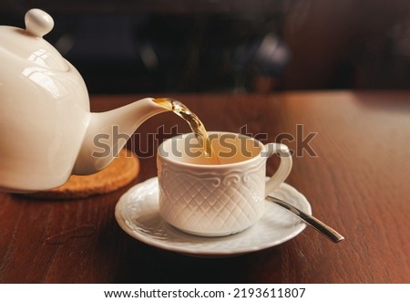 White cup with teapot. The hand holding the teapot pours tea into a pot standing on saucer in soft focus on naturally blurred background. Coffee, tea house, bokeh lights. The concept of a cozy