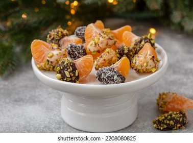 White and dark Chocolate Dipped Clementine Slices sprinkled with chopped nuts and coconut flakes served on white ceramic cake stand. Festive citrus dessert. Horizontal, selective focus.