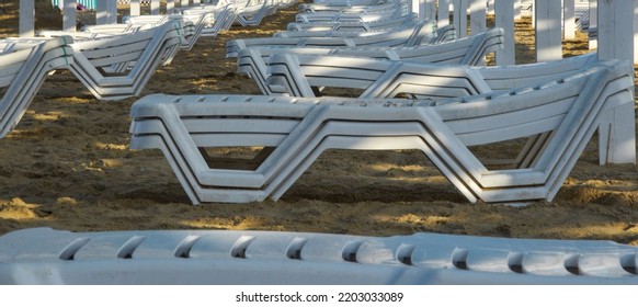 White Deck Chairs Stand In Even Rows On The Sandy Shore. An Awning Is Stretched Over The Sun Loungers, It Keeps A Shadow Over The Vacationers. White, Plastic Deck Chairs On The Coastal Strip.