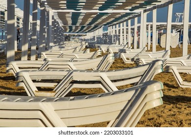 White Deck Chairs Stand In Even Rows On The Sandy Shore. An Awning Is Stretched Over The Sun Loungers, It Keeps A Shadow Over The Vacationers. White, Plastic Deck Chairs On The Coastal Strip.