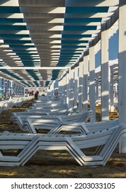 White Deck Chairs Stand In Even Rows On The Sandy Shore. An Awning Is Stretched Over The Sun Loungers, It Keeps A Shadow Over The Vacationers. White, Plastic Deck Chairs On The Coastal Strip.