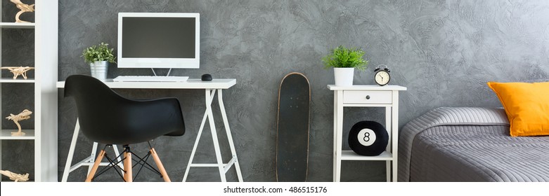 White Desk With Computer By The Grey Wall In Boy's Bedroom. Next To It Skateboard, Nightstand And Big Bed