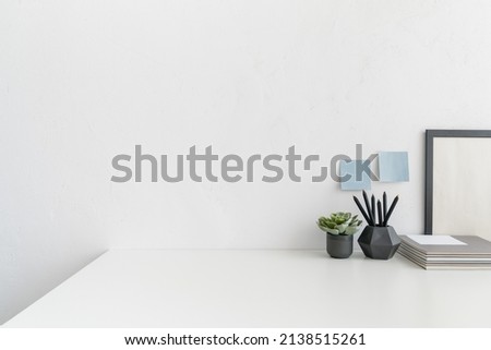 White desk, with empty space. Workspace with design supplies, flowers in a vase against white wall. 