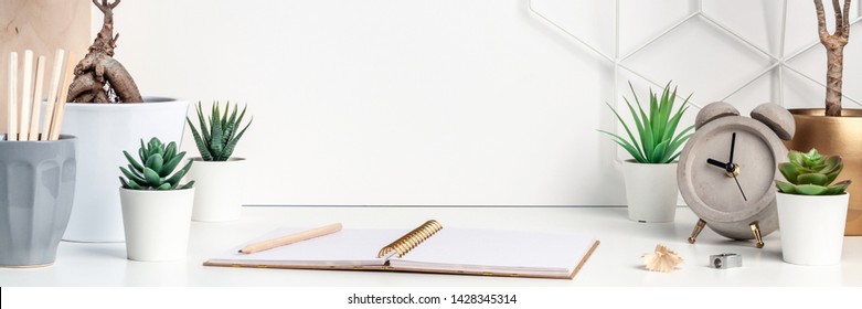 White desk at an empty wall with a geometric pattern. Copy space. Green succulents, bonsai, wooden stand, open notebook and gray concrete clock. Bright composition. Panoramic real photo - Powered by Shutterstock