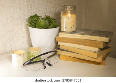 A white desk features a stack of books, a lit jar candle, two decorative holders, a potted succulent, and clear safety glasses.