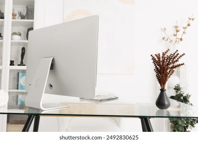 A white desktop computer sits on a glass desk with a black metal frame. There are dried flowers in a black vase to the right of the computer, and a bookshelf in the background.