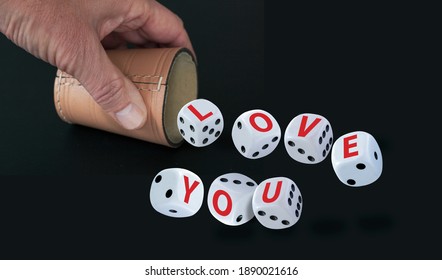 White Dice With Black Eyes Numbers Spelling LOVE YOU  In Red Letters Rolling Out From A Raffle Cup Held By A Hand Isolated On Black Background. Love As A Game Concept.