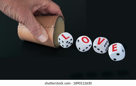 White Dice With Black Eyes Numbers Spelling LOVE  In Red Letters Rolling Out From A Raffle Cup Held By A Hand Isolated On Black Background. Love As A Game Concept.