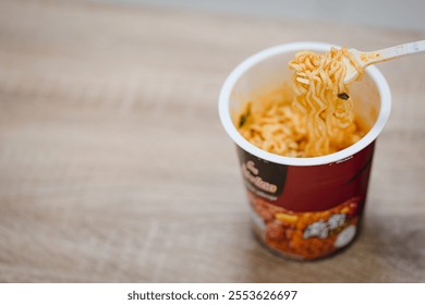 white disposable plastic fork with cooked instant noodles over blurred disposable paper cup with aluminum foil lid on wooden background. Eating behavior, effects of instant noodles on human health.