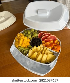 White divided serving tray with ice compartment filled with assorted fresh sliced fruits on wooden table with open lid