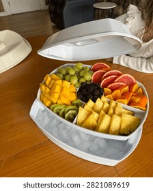 White divided serving tray with ice compartment filled with assorted fresh sliced fruits on wooden table with open lid