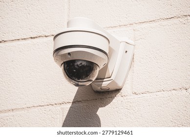 A white dome security camera is attached to a rough exterior wall, casting a distinct shadow