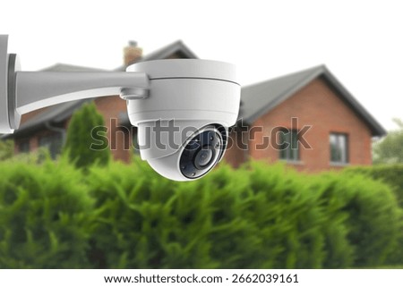 A white dome security camera is prominently displayed in the foreground, mounted on a wall. In the blurred background, a brick house with a grey roof is visible, surrounded by lush green bushes.