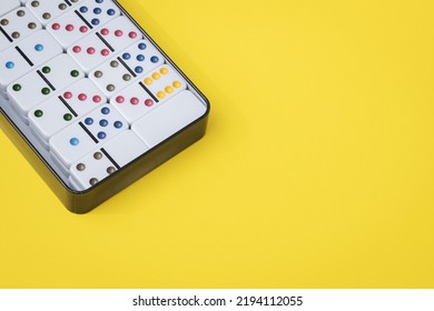 White Dominoes With Colored Dots In A Metal Box Lie In The Upper Left Corner On A Yellow Background With Copy Space On The Right, Flat Lay Close Up. Summer Board Game Concept.