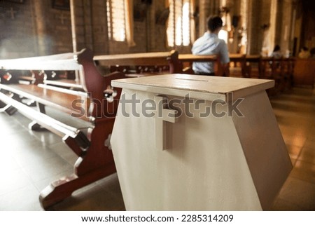 White donation box with a cross on it at a Christian church.  Collection box for generous offertory standing among pews at a Catholic temple