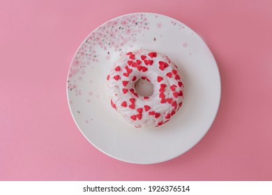 White donut sprinkled with small pink hearts placed on a beautiful white plate with pink flowers. Pink background.