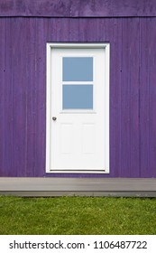 A white door with two windows is centered on a wooden exterior wall, painted purple. A small gray-brown porch separates the building from a thick strip of green grass. A simple colorful background.