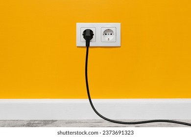 White double outlet installed on yellow wall with inserted black electrical plug, front view.