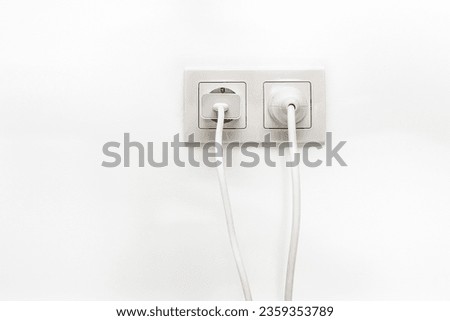 White double outlet installed on the white wall with inserted phone adapter and a white electrical plug, front view.