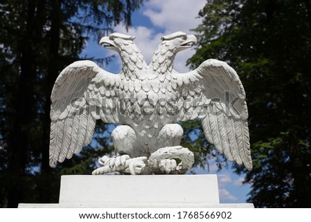 White doubleheaded metal eagle, looking in different directions, against the background of trees and blue sky with clouds.