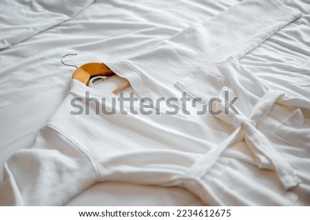 A white down bathrobe decorated,soft and comfortable fabric, hung on hanger on a bed with white linens.It is a set for hotel guests to use while bathing or wear bathing suit.Time to travel, journey,