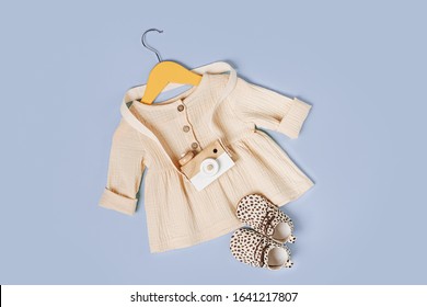 White Dress On Hanger With Toy Camera And Shoes. Set Of Baby Clothes And Accessories For Spring, Autumn Or Summer On Light Blue Background. Fashion Kids Outfit. Flat Lay, Top View