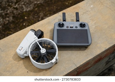 A white drone with a circular propeller system rests beside its remote control on a weathered concrete surface under natural daylight showing technical components ready for operation.