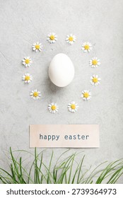 White Easter egg framed by chamomile daisies with Happy Easter card and fresh green grass on light concrete background. Minimal spring flat lay with copy space.