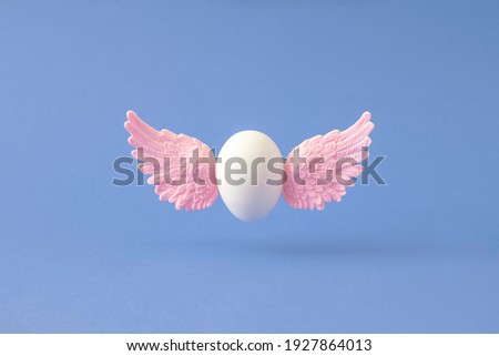 White Easter egg with pink angel wings flying against blue background. Creative resurrection concept.