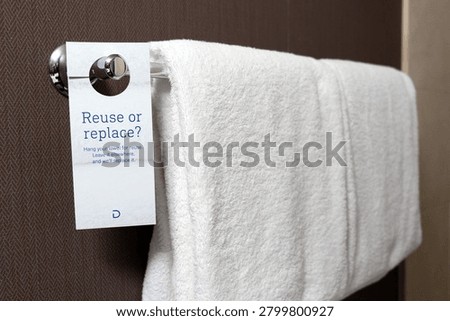 A white eco friendly reuse or replace sign hangs on a chrome rack next to a folded white bath towel against a textured brown wall, encouraging hotel guests to save water for environmental friendly