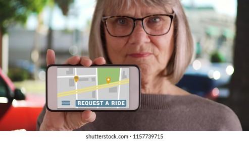 White Elderly Female Outside On Sidewalk Holding Phone With Rideshare App Open