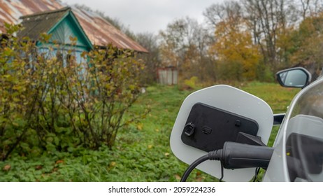 White Electric Car Is Charged In The Village. Outside The City The Countryside. Private Electric Car Charger. Electric Charge In A Village House