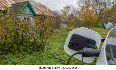 White Electric Car Is Charged In The Village. Outside The City The Countryside. Private Electric Car Charger. Electric Charge In A Village House