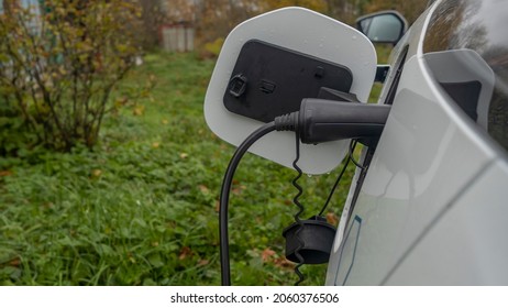 White Electric Car Is Charged In The Village. Outside The City The Countryside. Private Electric Car Charger. Electric Charge In A Village House