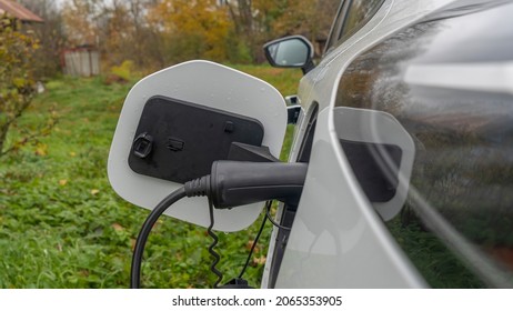 White Electric Car Is Charged In The Village. Outside The City The Countryside. Private Electric Car Charger. Electric Charge In A Village House