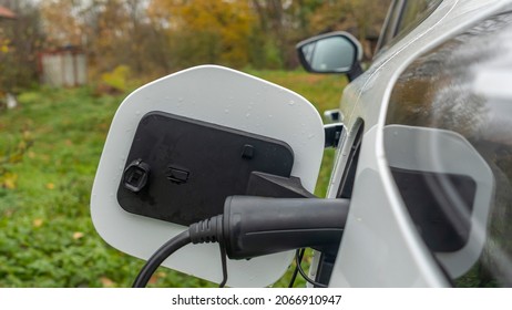 White Electric Car Is Charged In The Village. Outside The City The Countryside. Private Electric Car Charger. Electric Charge In A Village House