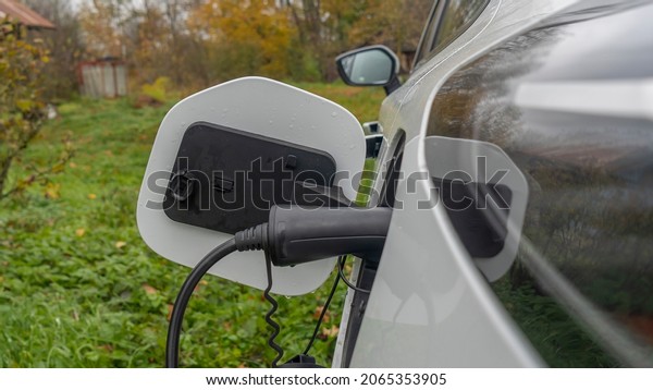 White electric car is charged in the\
village. Outside the city the countryside. Private electric car\
charger. Electric charge in a village\
house
