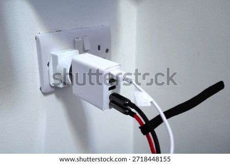 A white electrical adapter is plugged into a wall socket, connected through a power converter. Three USB ports on the adapter are in use, with white, black and red cables attached. The wall plate shows a UK-style socket.