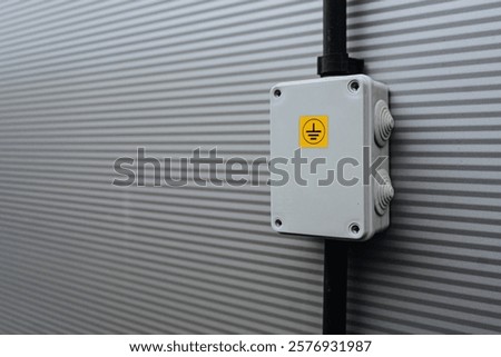 
A white electrical junction box with a yellow grounding symbol is securely mounted on a corrugated gray metal wall. Black wiring connects to the box, creating an industrial, clean, and organized look