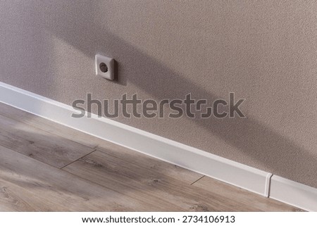 A white electrical socket mounted on a beige textured wall above a white skirting board, adjacent to light wooden flooring. Clean, minimalist interior detail.