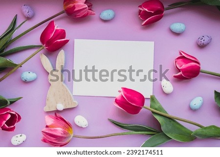 White empty envelope copy space Easter eggs and wooden bunnies with pink tulips on violet pastel background. Greeting card Postcard. Flat lay composition top view. Decoration minimalist modern design