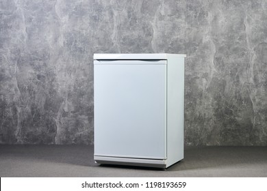 White Empty Mini Fridge Or Mini Bar Against Gray Textured Wall Background. International Exhibition Furniture Elements In Large Warehouse Interior.