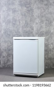 White Empty Mini Fridge Or Mini Bar Against Gray Textured Wall Background. International Exhibition Furniture Elements In Large Warehouse Interior.